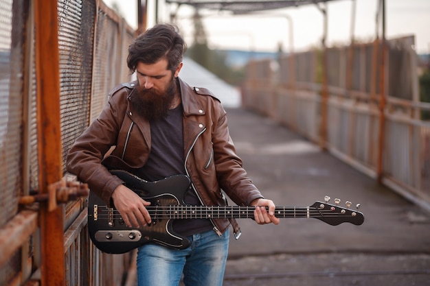 Bearded rocker with gray hair in a brown leather jacket and blue jeans stands and holds a black electric guitar against the background of a road