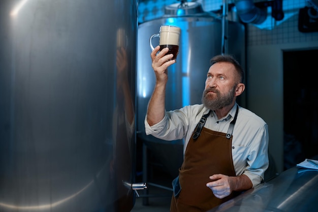 Bearded senior brewer checking beer quality during brewing technology process. Mature brew master looking at foam level in glass