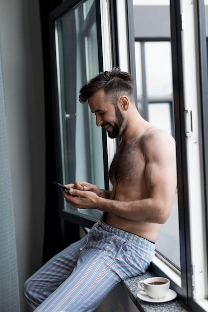 Bearded shirtless man using smartphone while sitting on windowsill and drinking coffee at home