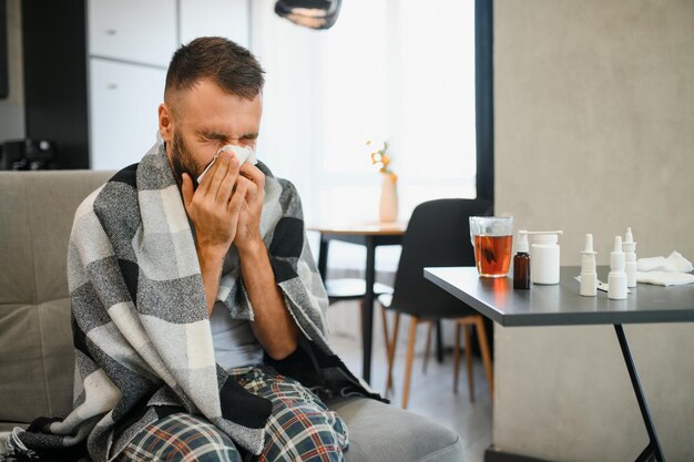 Photo bearded sick sad man sits on couch at home suffers from runny nose flu disease coronavirus pandemic covid epidemic sneezes