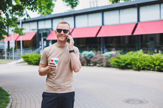 A bearded smiling stylish man in white shirt and sunglasses standing on the streets of the city and calling on mobile phone