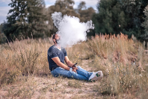 Bearded smoker blowing a lot of smoke using vape electronic cigarette in nature. Electronic cigarette.