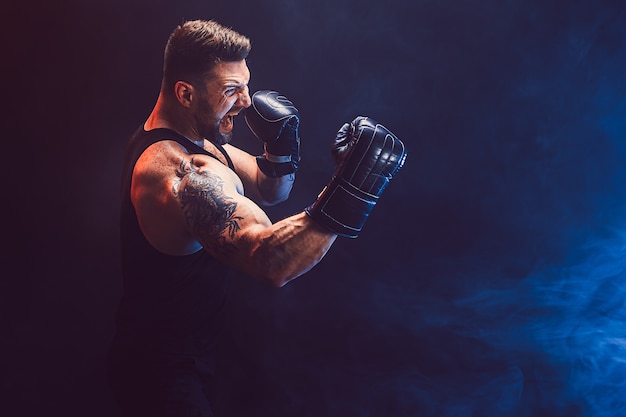 Photo bearded tattooed sportsman muay thai boxer in black undershirt and boxing gloves fighting on dark wall with smoke. sport concept.