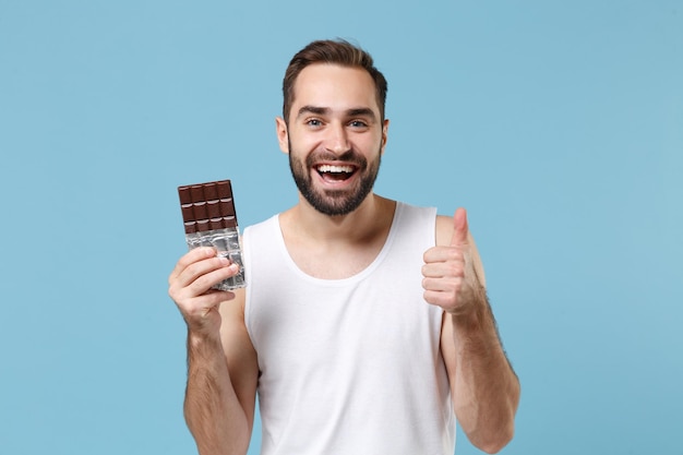 Bearded young man 20s years old in white shirt hold in hand chocolate bar isolated on blue pastel wall background, studio portrait. Skin care healthcare cosmetic procedures concept. Mock up copy space