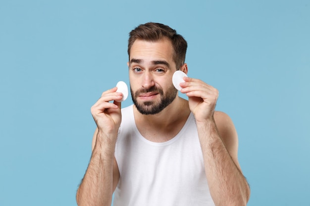 Bearded young man 20s years old in white shirt hold in hand cotton pad isolated on blue pastel wall background, studio portrait. Skin care healthcare cosmetic procedures concept. Mock up copy space.