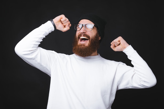 Bearded young man is screaming of excitement with both hands up on black background.