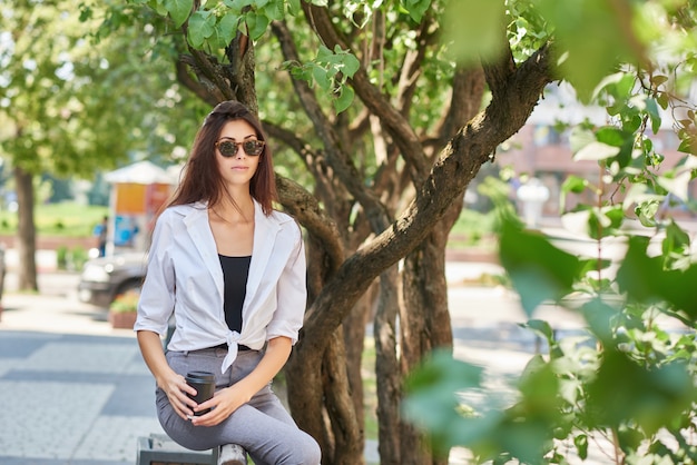 Beatiful young student sitting on wooden bench, keeping paper coffee can.