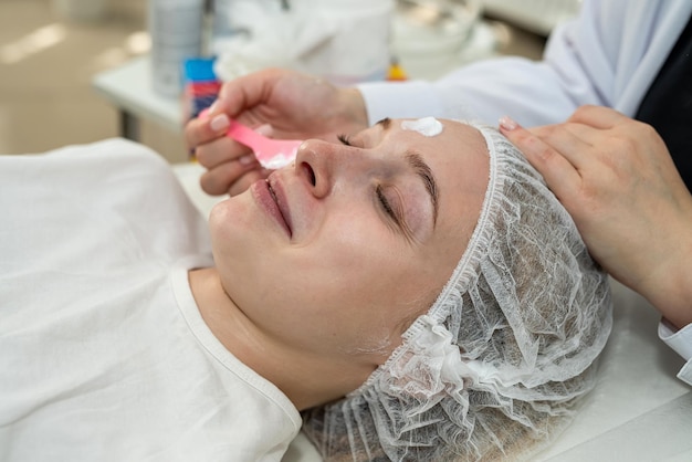 Beautician applies cleansing and moisturizing white cream with help of brush on clients face