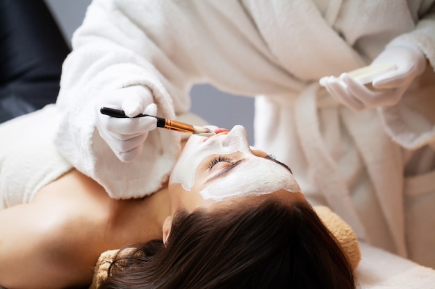 Beautician applies a mask on the face of a woman lying on a couch in a spa