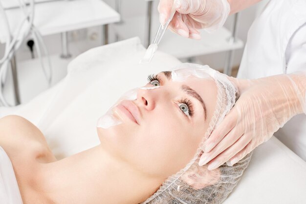 Beautician applies vitamins cream mask on woman face for rehydrate face skin in beauty salon