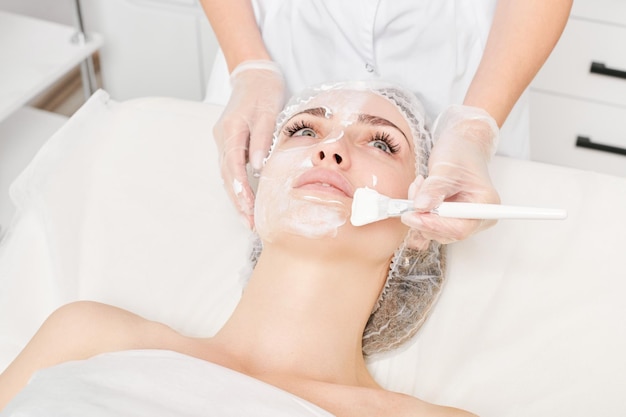 Beautician applies vitamins cream mask on woman face for rehydrate face skin in beauty salon