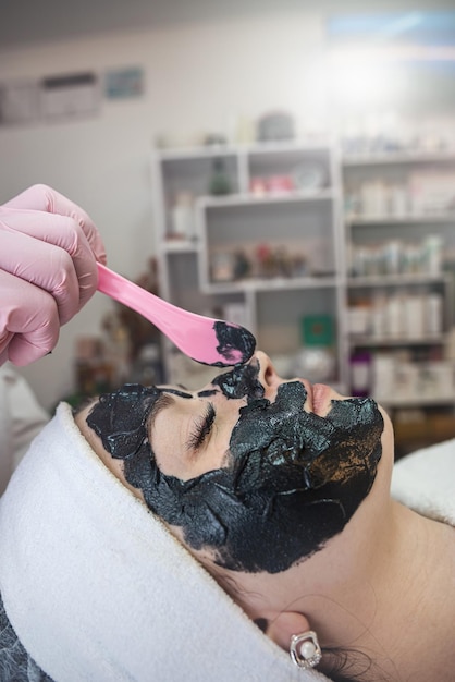 Beautician applying black mask at young pretty woman face in spa salon