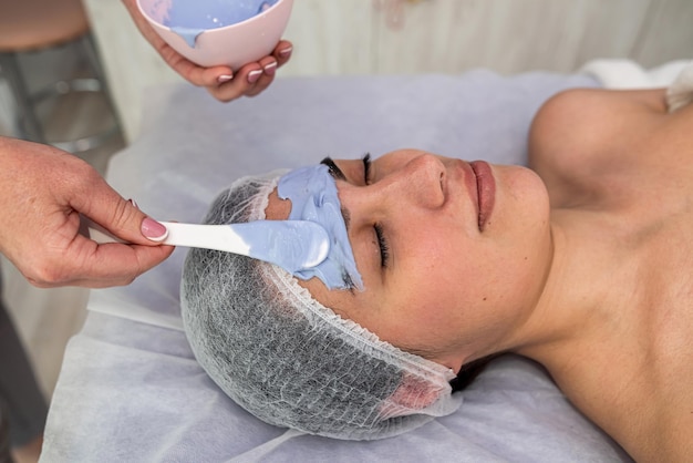 Beautician applying blue facial cleansing cream mask on female face clients