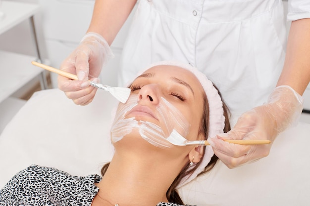 Beautician applying cosmetic cream mask on woman face for rejuvenation procedure in beauty salon