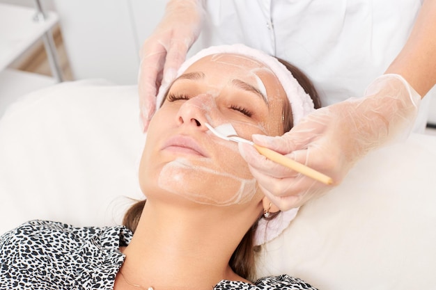 Beautician applying cosmetic cream mask on woman face for rejuvenation procedure in beauty salon