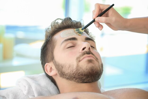 Beautician applying scrub onto young man's face in spa salon