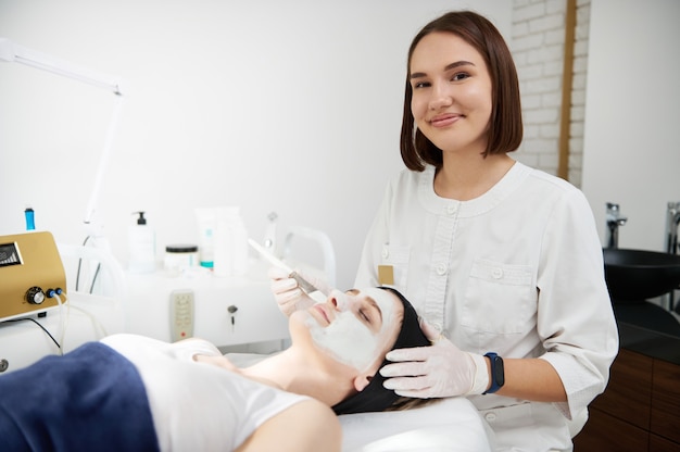 Beautician doctor applying beauty moisturizing mask to face of blonde woman lying on massage table in beauty spa clinic. Professional skin care concept in beauty salons with modern equipment