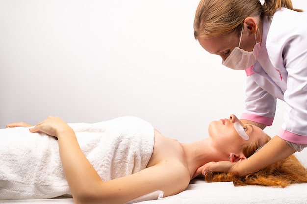 The beautician gives the woman a professional face neck massage photo on white wall