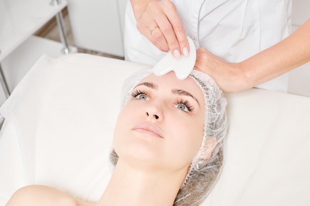 Beautician making facial massage with Gua Sha stone of woman face skin for lymphatic drainage