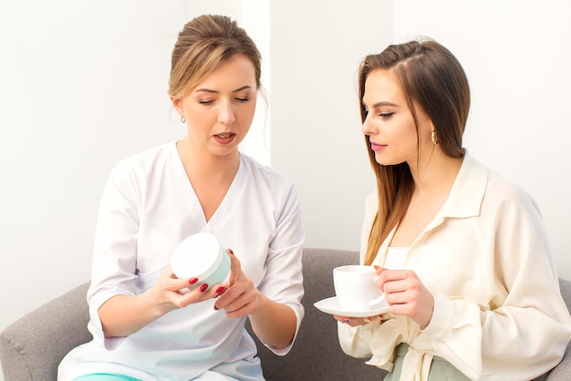 Beautician offering product for the young woman holding a white plastic jar with a cream sitting on the sofa