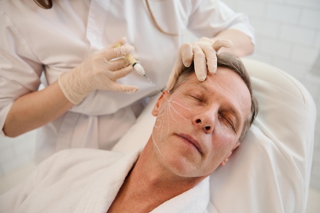 Beautician performs thread lifting according to the lines drawn on the mature handsome man's face. Rejuvenation, anti aging therapy concept for men in a modern spa clinic. Men's injection cosmetology