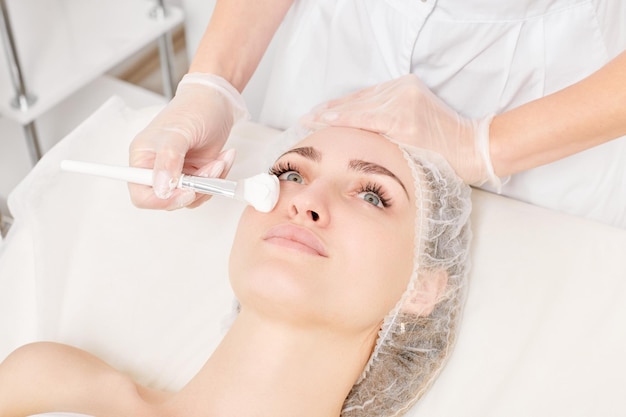 Beautician puts cream mask on woman face for rejuvenation face skin procedure in beauty salon