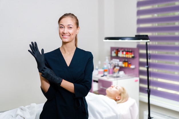 Beautician stands puts black gloves on  hand and smiles.