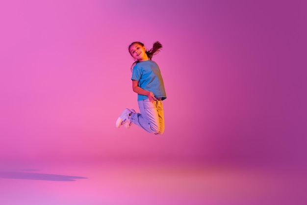 Beautiful active little girl kid jumping dancing isolated over pink background in neon action dance