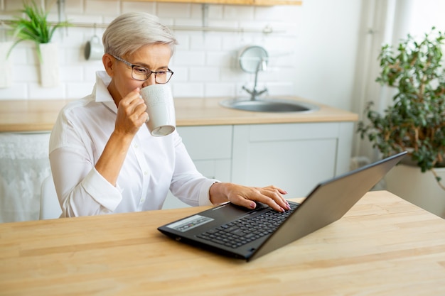 beautiful adult woman in glasses at the table with laptop at home with a mug