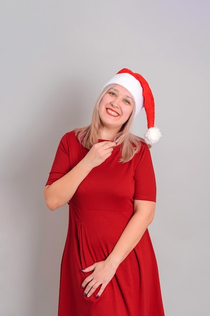 A beautiful adult woman in a Santa hat and a red dress poses on a gray background vertical photo