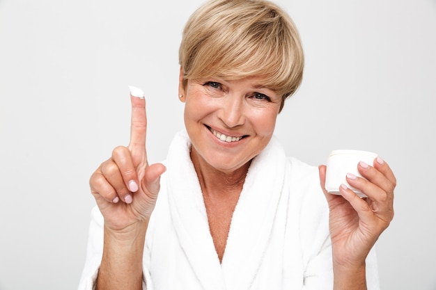  beautiful adult woman with short blond hair wearing white housecoat holding face cream jar isolated over white wall