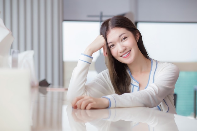 Beautiful adult young age Asian professional woman with long hair in white shirt looks at the camera her smiling happily while she sits on chair in workplace office with glassed building as a backgrou