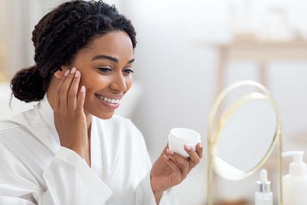 Beautiful African American Female Looking At Mirror And Applying Moisturising Face Cream