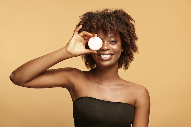 Beautiful african american girl covers the eye with a jar of cream and smiling