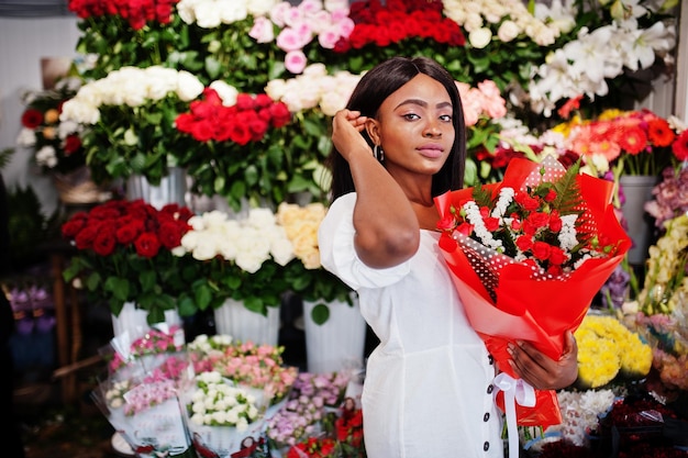 Beautiful african american girl in tender white dress with bouquet flowers in hands standing against floral background in flower shopBlack female florist