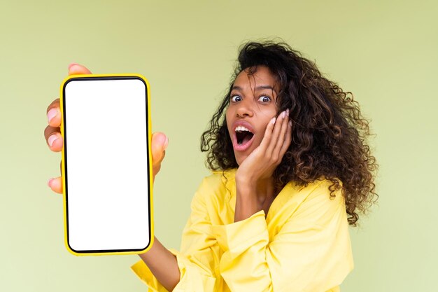 Beautiful african american woman in casual shirt on green background holds a phone with a blank white screen