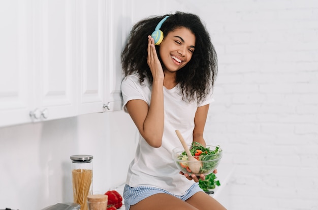 Beautiful African American woman cooking vegetarian dinner in the kitchen. Healthy lifestyle concept. Happy emotional girl holding bawl with fresh salad, listening music at home, laughing