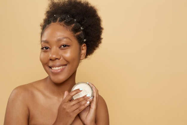 Beautiful african american woman holding jar of moisturizer or mask on beige background with copy space Skin care anti aging