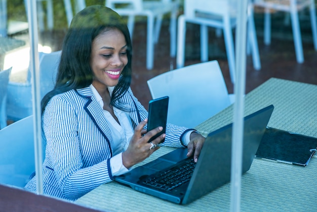 Beautiful African-American woman using a laptop and looking at her cell phone behind a window