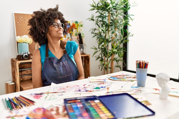 Beautiful african american woman with afro hair painting at art studio looking away to side with smile on face natural expression laughing confident