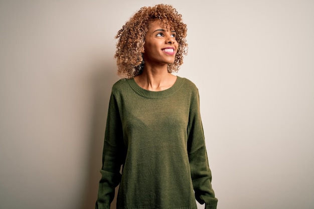 Beautiful african american woman with curly hair wearing casual sweater over white background looking away to side with smile on face natural expression Laughing confident