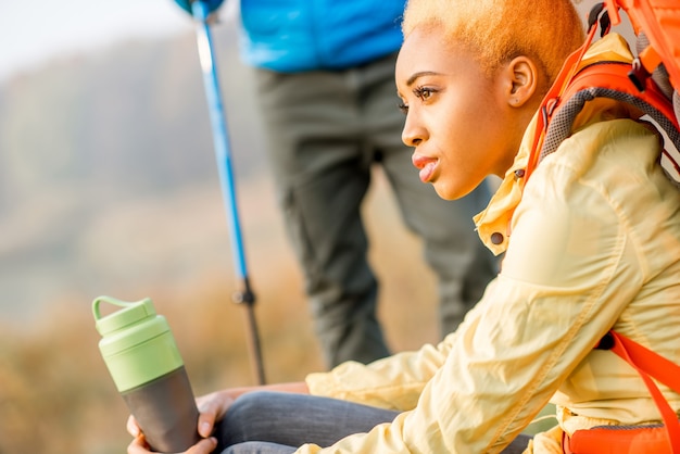 Beautiful african woman hiker in yellow jacket drinking water from the bottle sitting outdoors