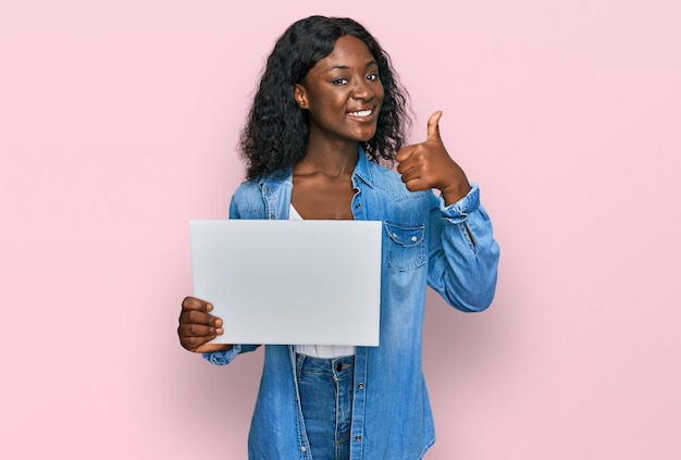 Beautiful african young woman holding blank empty banner smiling happy and positive, thumb up doing excellent and approval sign