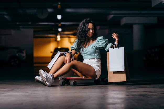 Photo beautiful afro american  sitting on a skateboard with shopping bags in the underground parking