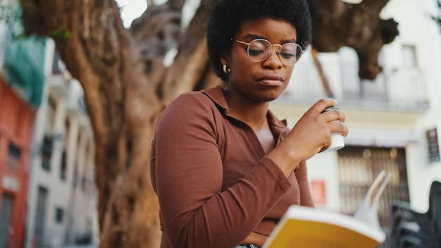 Beautiful Afro woman reading a book on the street wearing glass