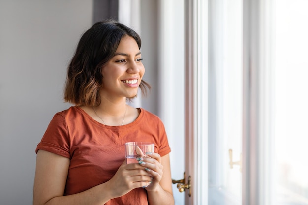 Beautiful Arab Woman With Glass Of Water In Hand Standing Near Window