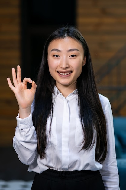 Beautiful Asian business woman smiling and showing OK sign standing in the hallway in the office