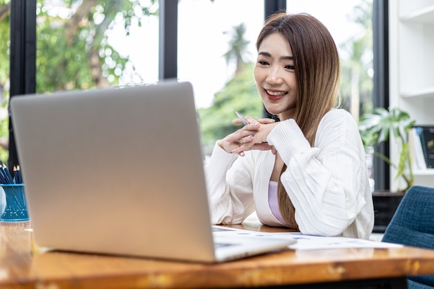 Beautiful Asian businesswoman sitting in her private office, she is talking to her partner via video call on her laptop, she is a female executive of a startup company. Concept of financial management