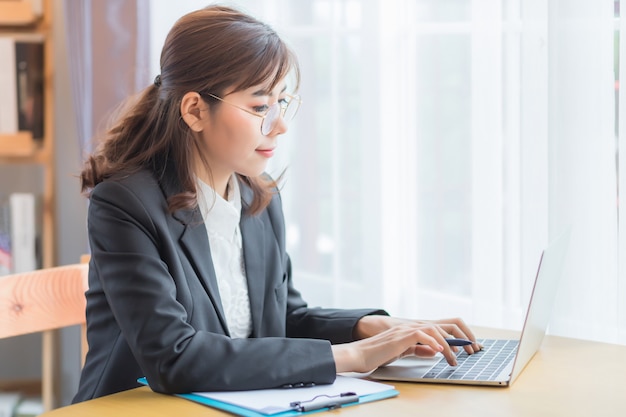 A beautiful Asian businesswoman. A Thai woman smiles and prints documents using a notebook on a wooden desk in the office in the morning.