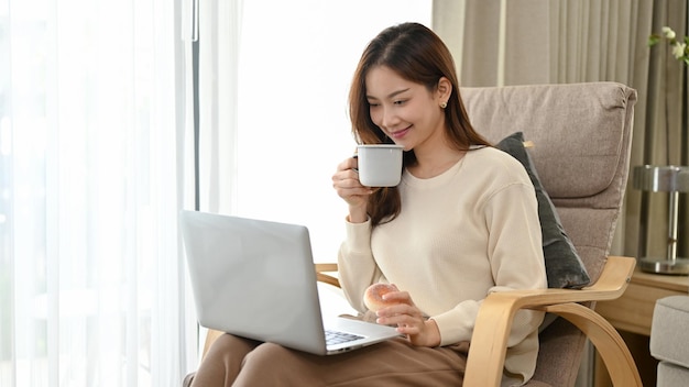 Beautiful Asian female or businesswoman working from home sipping coffee and using a laptop